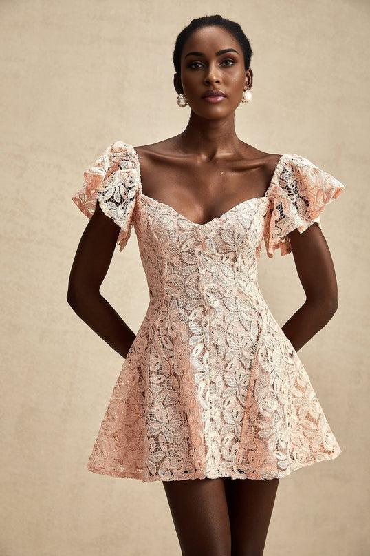 Young woman wearing a pink lace mini dress with short flutter sleeves standing in front of a plain background