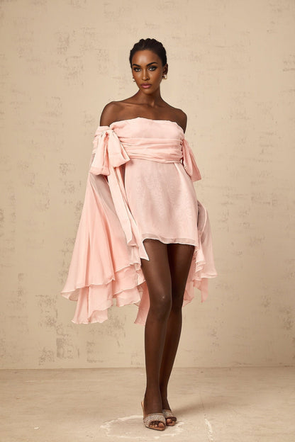Young woman wearing a pink mini dress with bow detail and draped fabric standing against a plain background