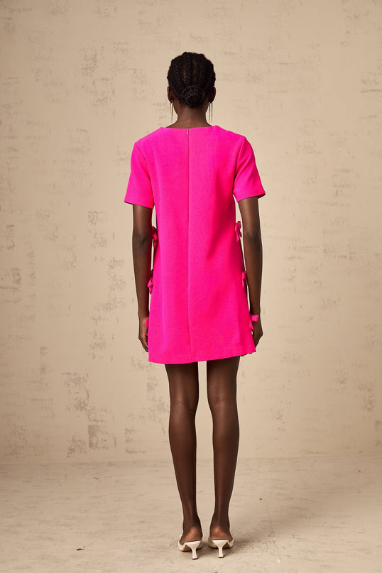 Young woman wearing a pink mini dress with bow detail on the front standing against a plain background