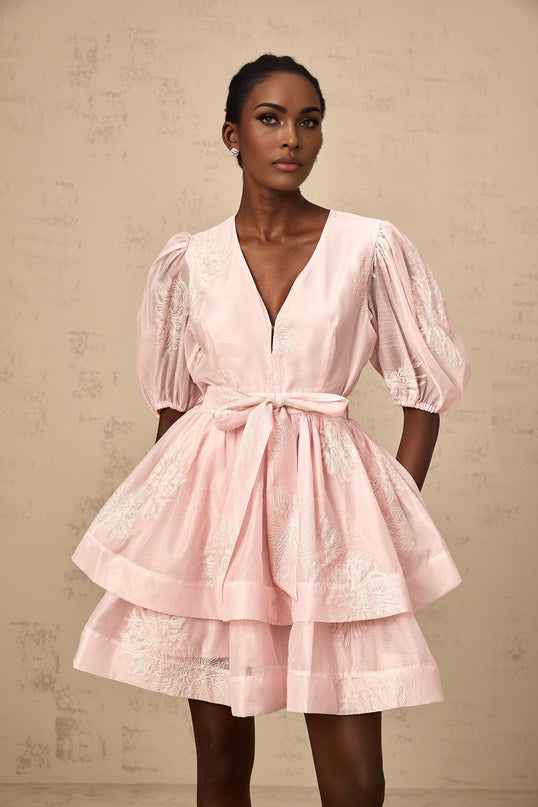 Young woman wearing a pink mini dress with floral embroidery and layered skirt against a plain background