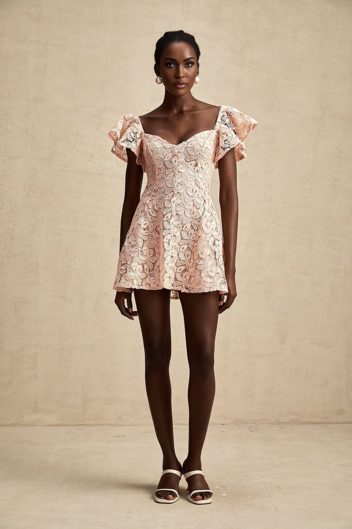 Young woman wearing a pink mini dress with flutter sleeves and lace detailing standing in a relaxed pose against a plain background