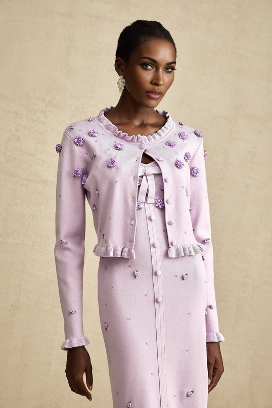 Young woman wearing a purple knitted cardigan and matching dress with faux flower embellishments standing against a plain background