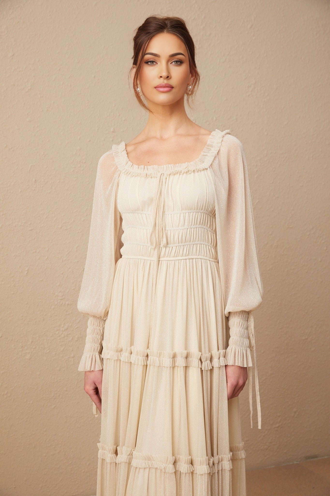 Young woman wearing a shimmering gold pleated midi dress with shirred detailing and short sleeves standing in front of a plain background