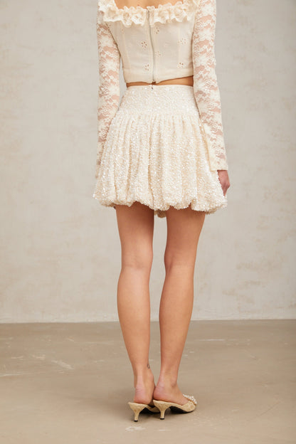 Young woman wearing a shimmering pleated mini skirt with sequin details standing against a plain background