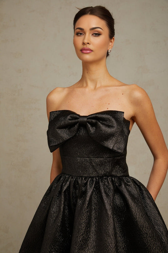 Young woman wearing a short black metallic dress with a bow detail standing against a plain background