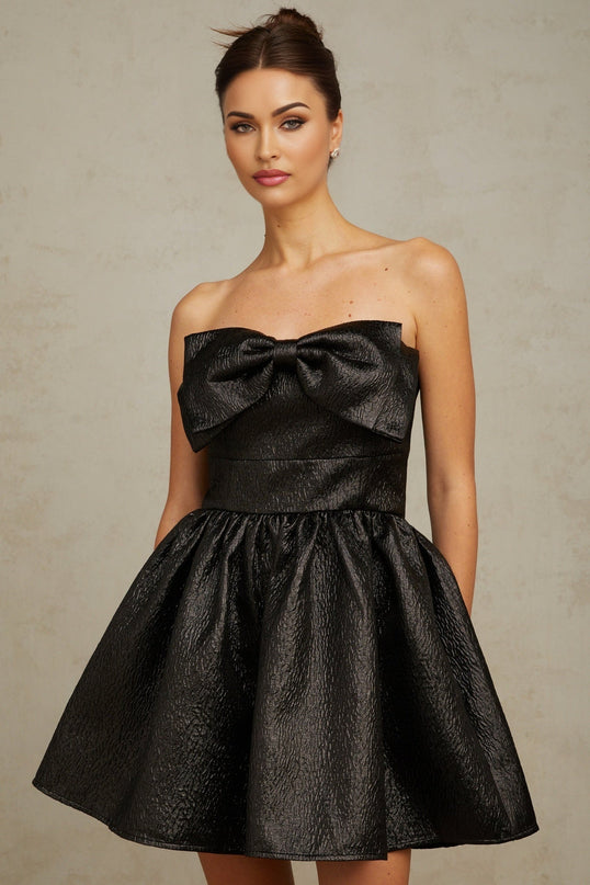 Young woman wearing a short black metallic dress with a bow detail standing against a plain background