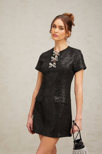 Young woman wearing a short black mini dress with glitter details standing against a plain background