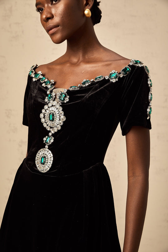 Young woman wearing a short black velvet dress with embellishments standing in front of a plain background
