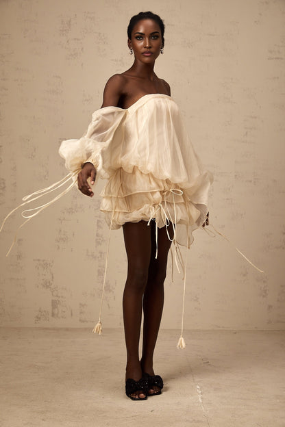Young woman wearing a short offshoulder dress with ruched fabric and layered skirt The dress has selftie straps and appears to be in a light color The model is posing against a plain background