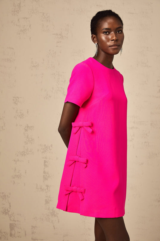 Young woman wearing a short pink dress with bow details and spaghetti straps standing against a plain background