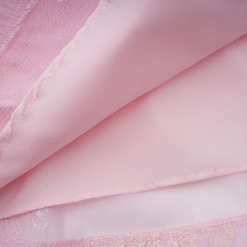Young woman wearing a short pink dress with floral embroidery and layered skirt standing in front of a light background