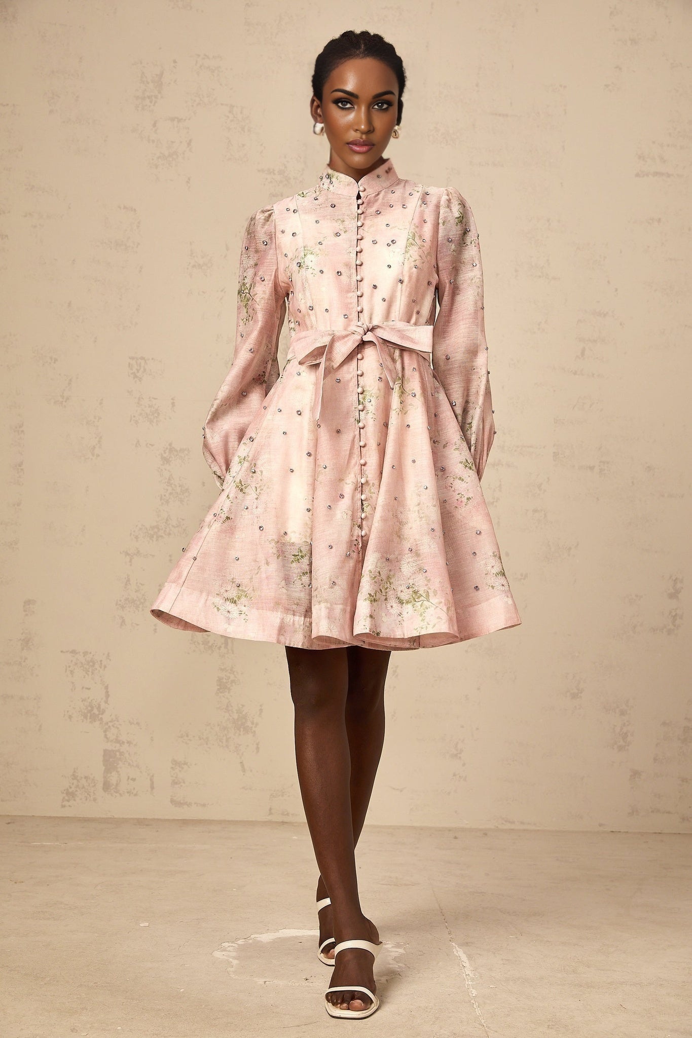 Young woman wearing a short pink dress with floral print and embellishments posing against a plain background