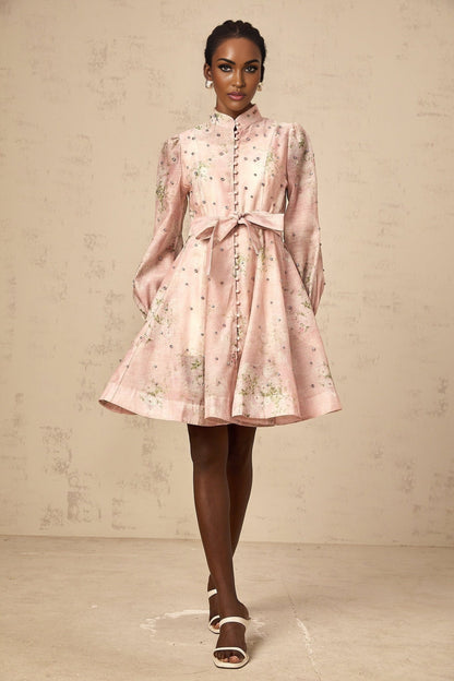 Young woman wearing a short pink dress with floral print and embellishments posing against a plain background