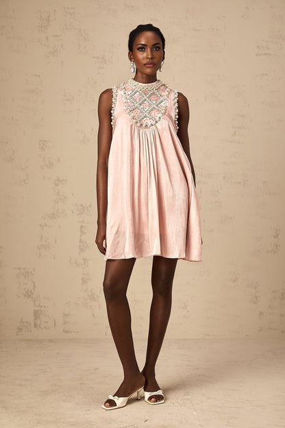 Young woman wearing a short pink dress with pleated skirt and embellished bodice standing in front of plain background