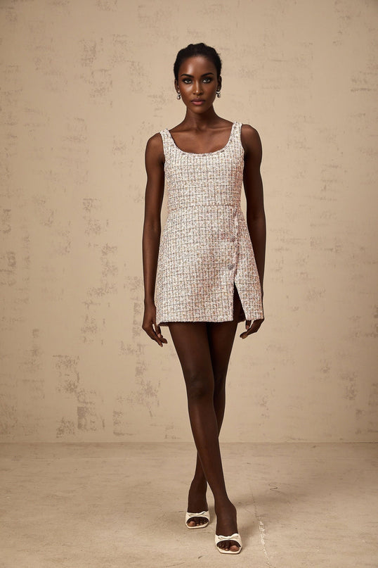 Young woman wearing a short pink tweed dress with embellishments standing against a plain background