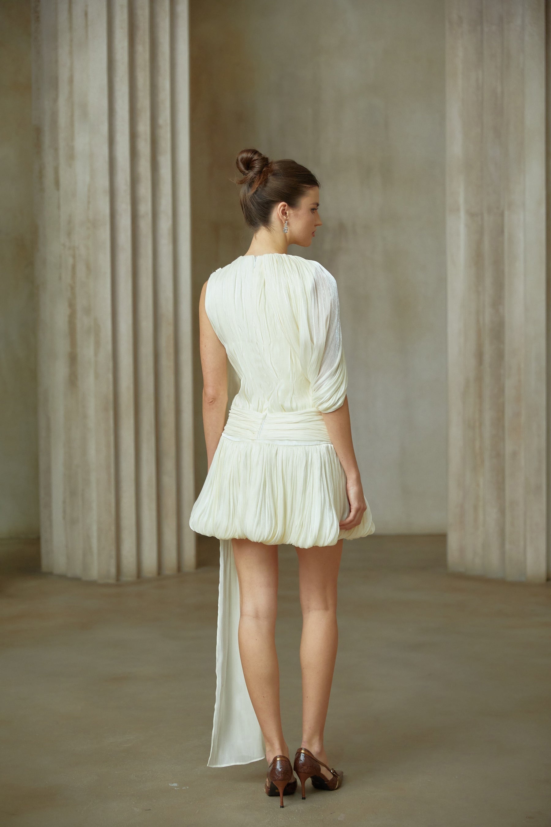 Young woman wearing a short pleated mini dress with a ribbon accent detail at the waist standing in a casual pose against a plain background