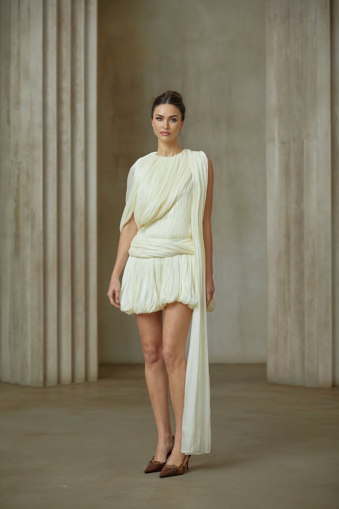 Young woman wearing a short pleated mini dress with a ribbon accent detail standing in a casual pose against a plain background