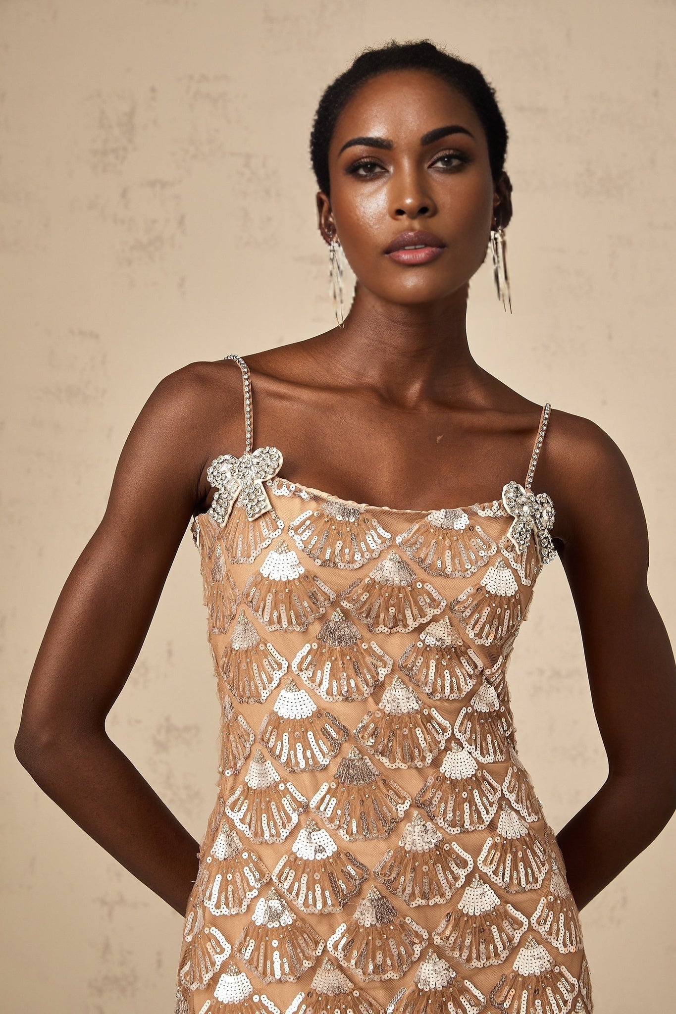 Young woman wearing a short shimmering silver dress with sequin embellishments standing against a plain background