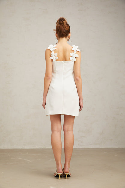 Young woman wearing a short white dress with floral embroidery standing in front of a plain background