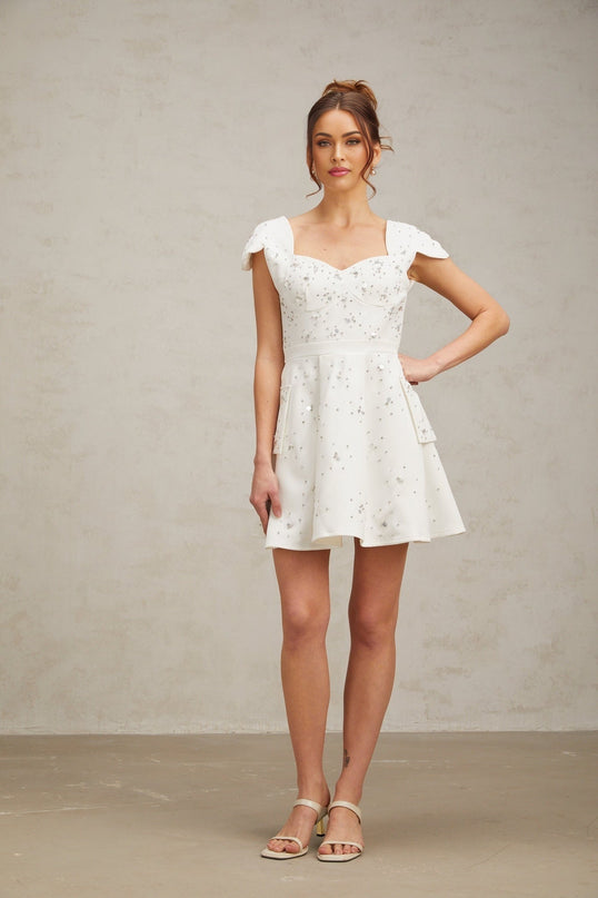 Young woman wearing a short white mini dress with flared skirt and embellished details standing against plain background