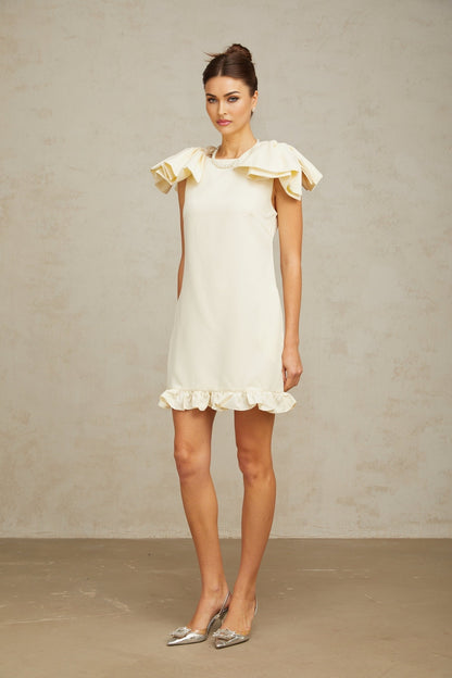 Young woman wearing a short white mini dress with ruffled details and a bow accent standing against a plain background