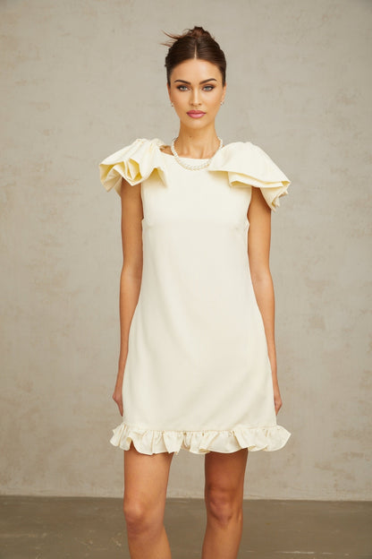 Young woman wearing a short white mini dress with ruffled details and a large bow accent standing against a plain background
