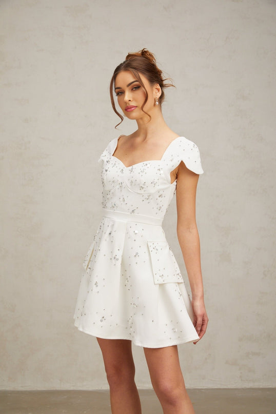 Young woman wearing a white flared mini dress with embellishments standing against a plain background