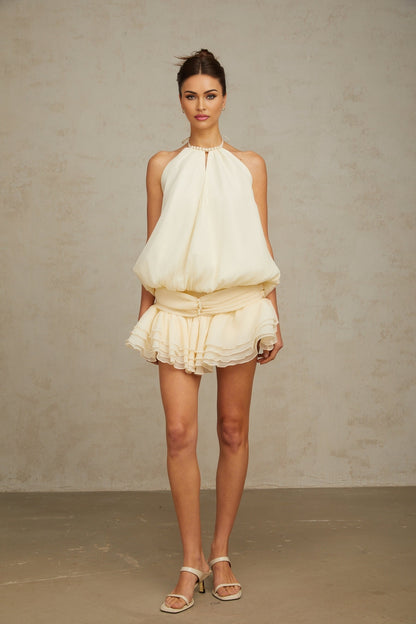 Young woman wearing a white halterneck mini dress adorned with faux pearls standing against a plain background