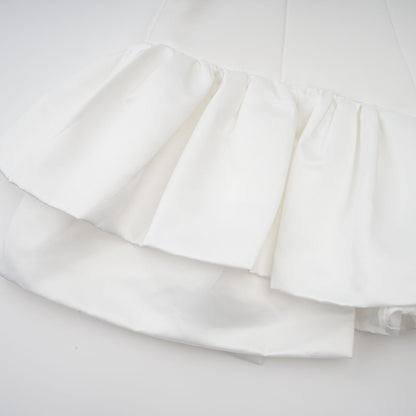 Young woman wearing a white halterneck mini dress with ruffled details standing against a plain background