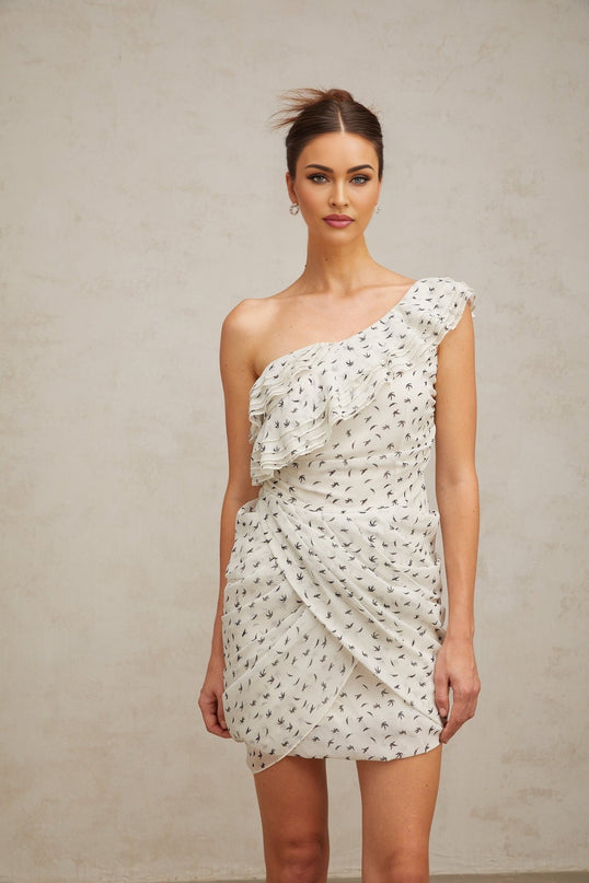 Young woman wearing a white mini dress with a pattern print and ruffled details standing against a plain background