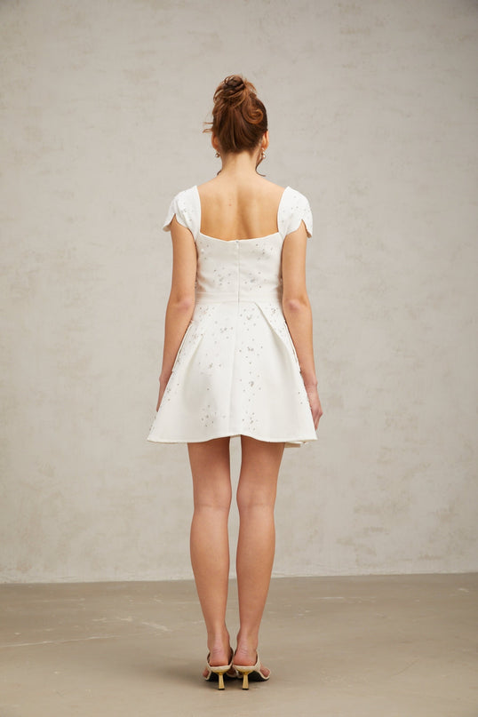 Young woman wearing a white mini dress with embellishments and a flared skirt standing against a plain background