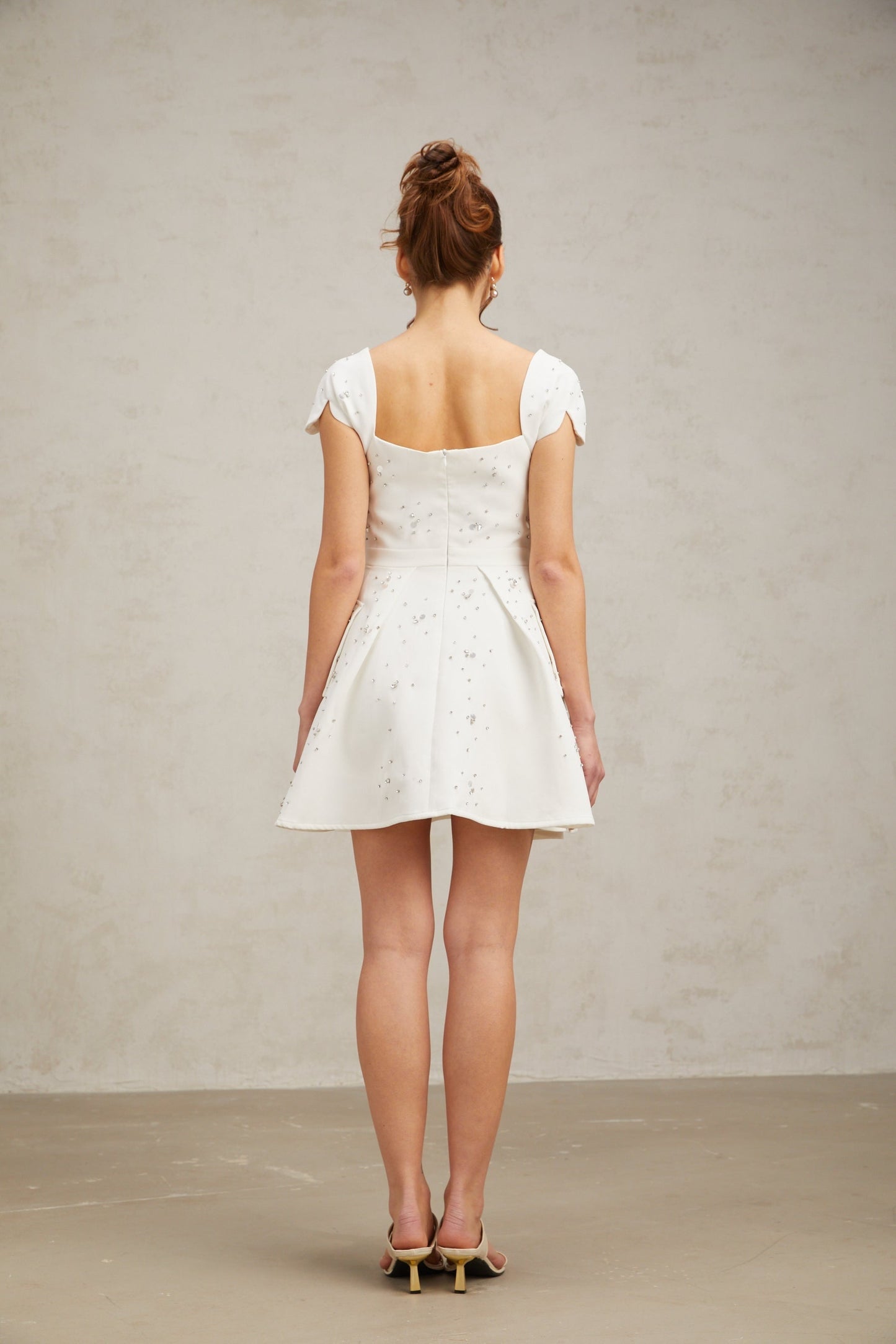 Young woman wearing a white mini dress with embellishments and a flared skirt standing against a plain background