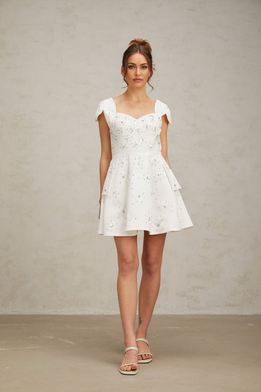 Young woman wearing a white mini dress with flared skirt and embellished details standing in a neutral setting