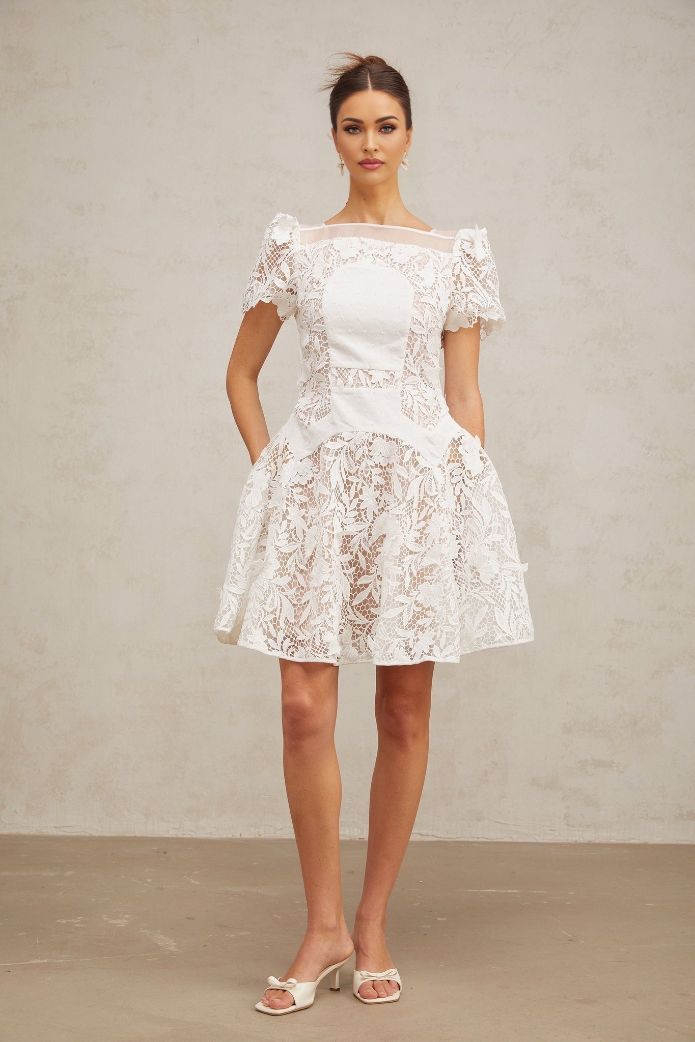 Young woman wearing a white mini dress with floral lace pattern and puff sleeves standing against a plain background
