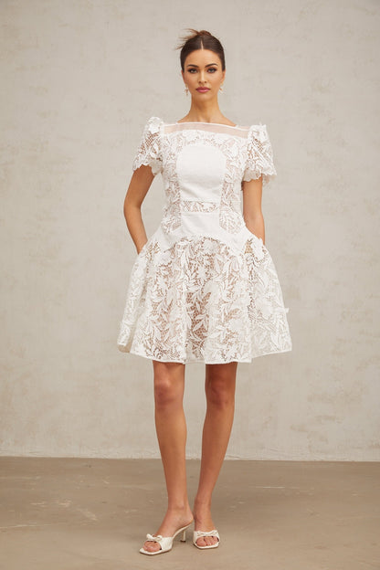 Young woman wearing a white mini dress with floral lace pattern and puff sleeves standing against a plain background