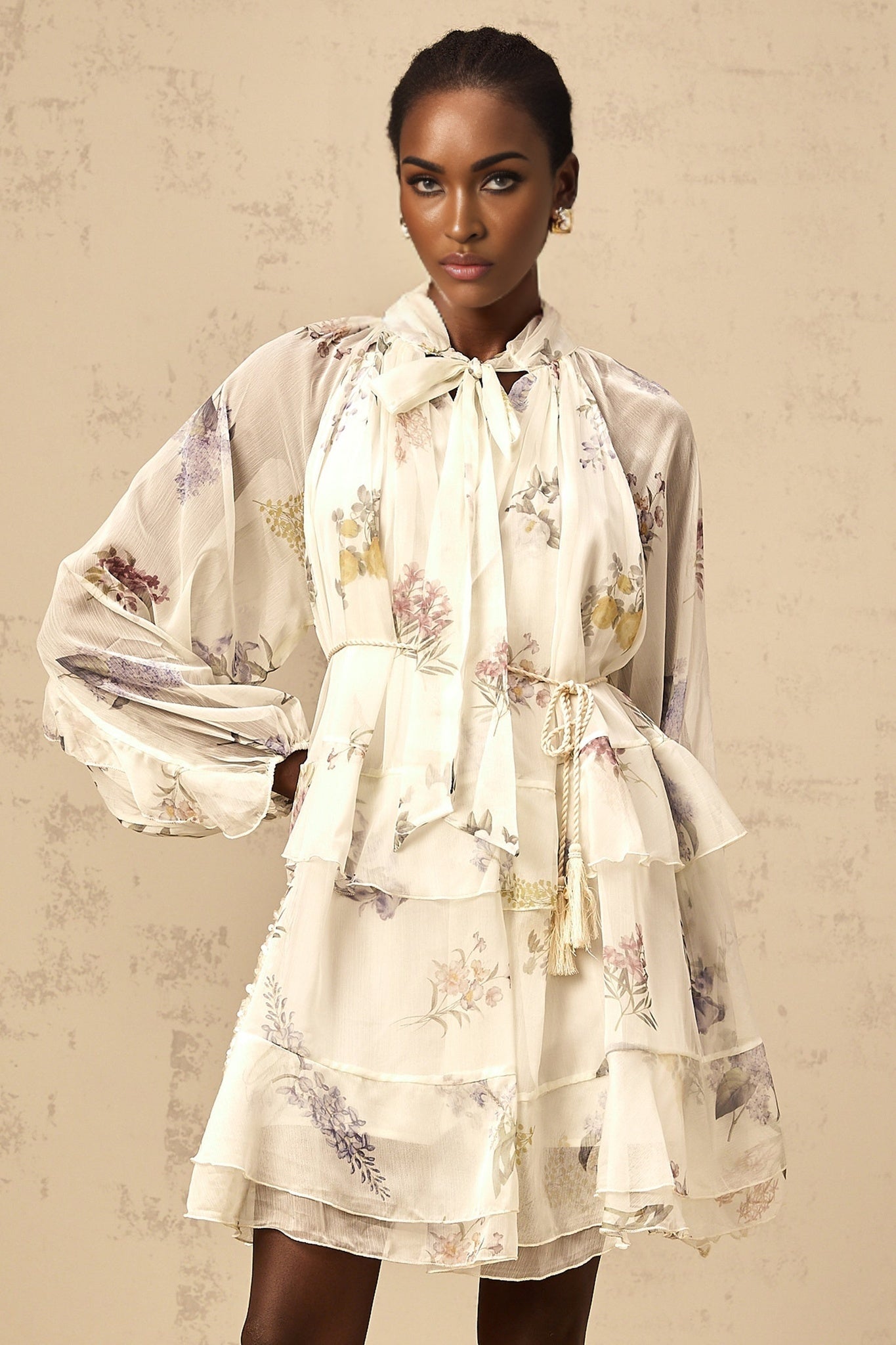 Young woman wearing a white mini dress with floral print and tiered chiffon skirt standing in front of a plain background
