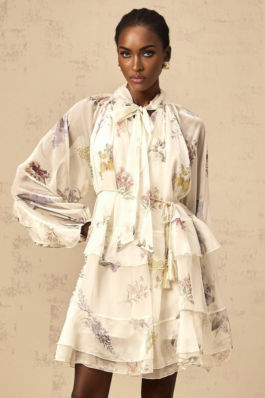 Young woman wearing a white mini dress with floral print and tiered chiffon skirt standing in front of a plain background