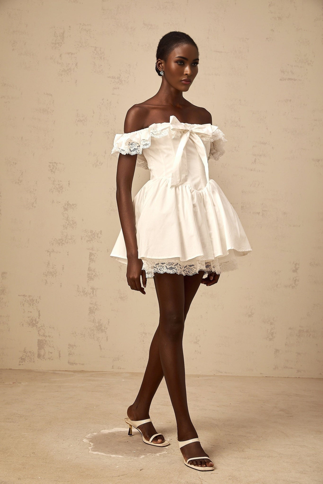 Young woman wearing a white mini dress with oversized bow and ruffled details standing against a plain background