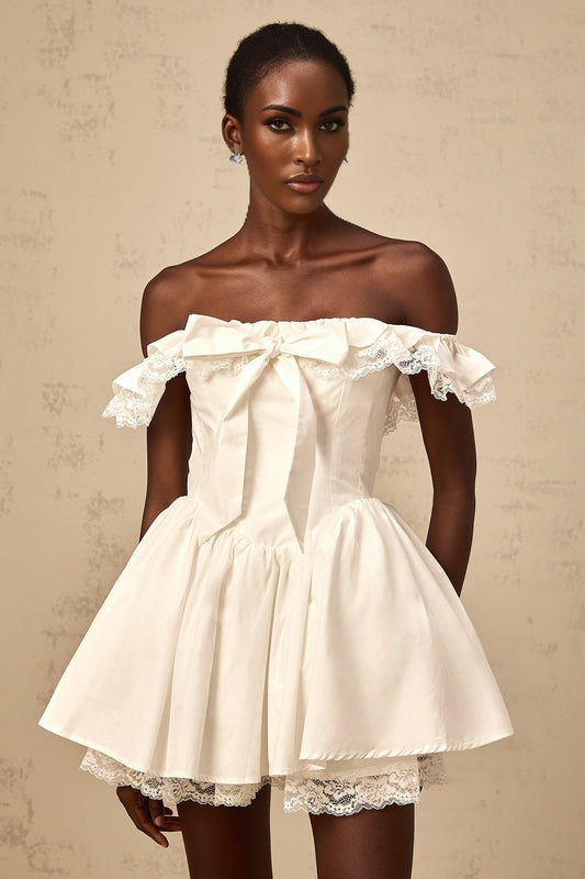 Young woman wearing a white mini dress with oversized bow and ruffled details standing against plain background