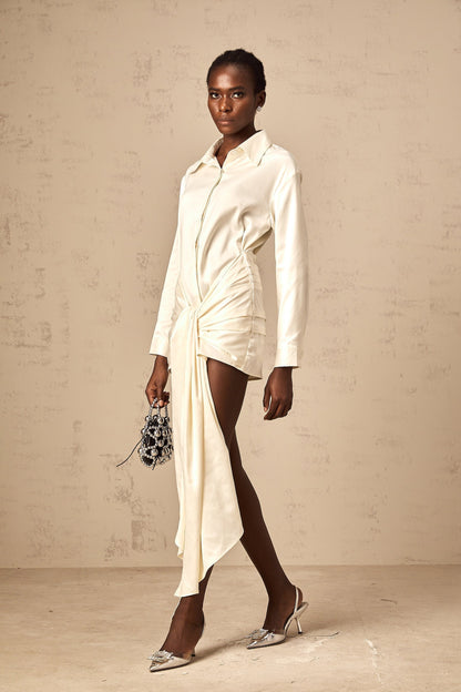 Young woman wearing a white mini shirt dress with ruched detailing and ribbon accents standing against a plain background