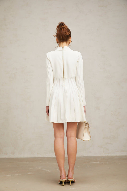 Young woman wearing a white pleated knit mini dress with short sleeves standing against a plain background