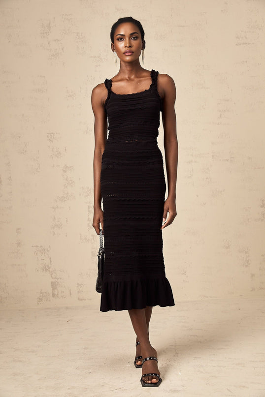 Young woman wearing black knitted matching set with ruffled top and skirt standing against plain background