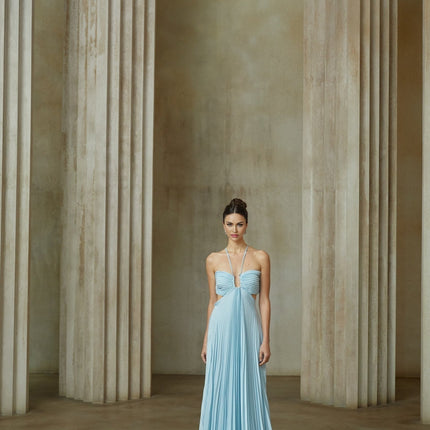 Young woman wearing long blue pleated maxi dress with cutout detail at waist standing against plain background