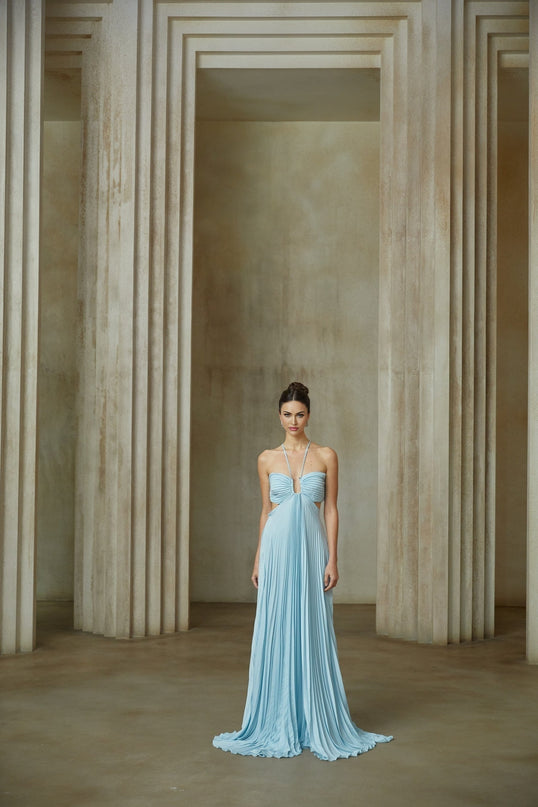 Young woman wearing long blue pleated maxi dress with cutout detail at waist standing against plain background