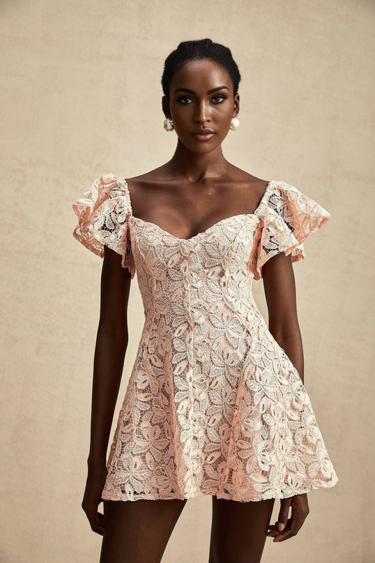 Young woman wearing pink lace mini dress with flutter sleeves posing against plain background